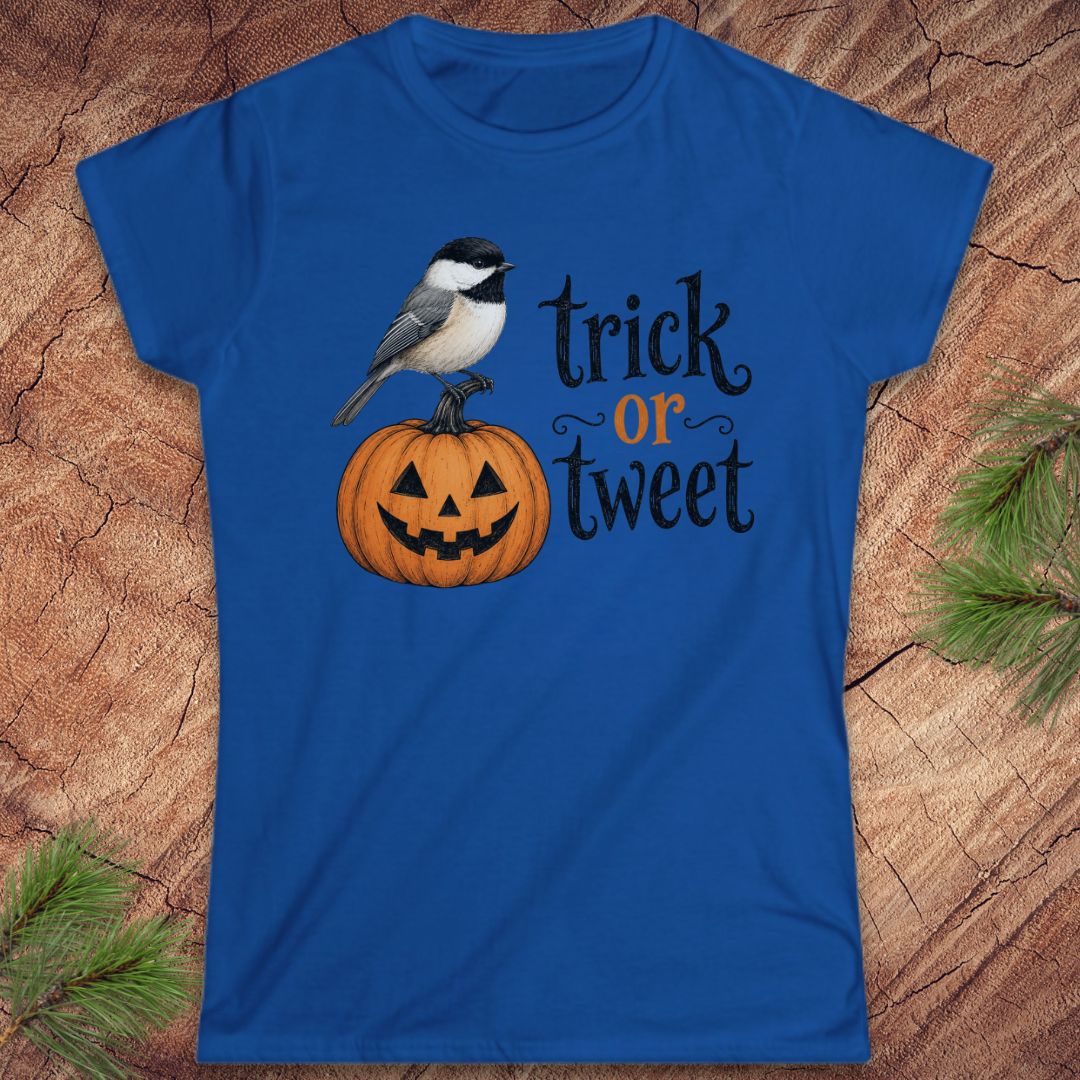 A royal blue short-sleeve t-shirt with a Halloween graphic featuring a bird perched on a jack-o'-lantern and the text 'trick or tweet' in black and orange, displayed flat on a wood background with pine branches.