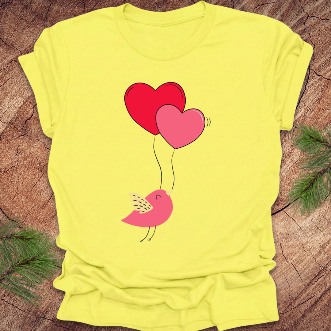 Yellow t-shirt with a pink bird holding heart-shaped balloons on a wooden surface.