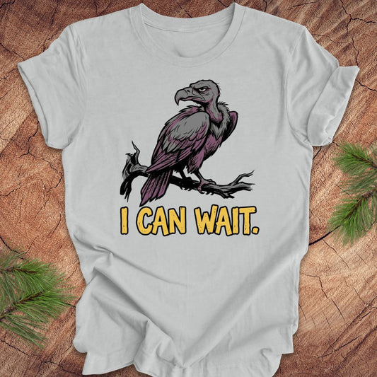 Silver t-shirt with a vulture graphic and 'I CAN WAIT.' text on a wooden surface.