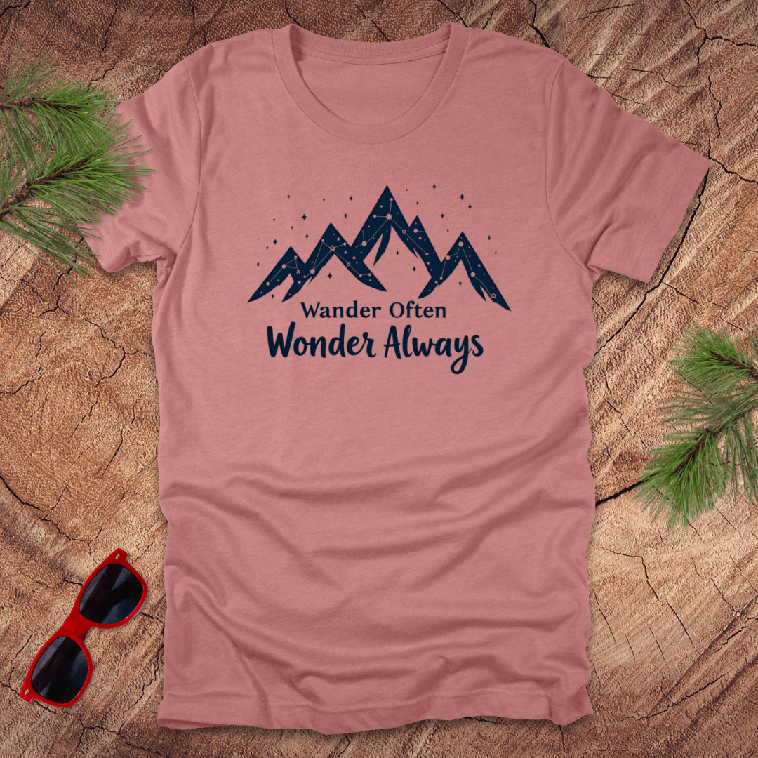 A mauve t-shirt with the text 'Wander Often Wonder Always' printed on the front, featuring a mountain graphic. The shirt is displayed on a wooden surface with pine needles and a pair of sunglasses.