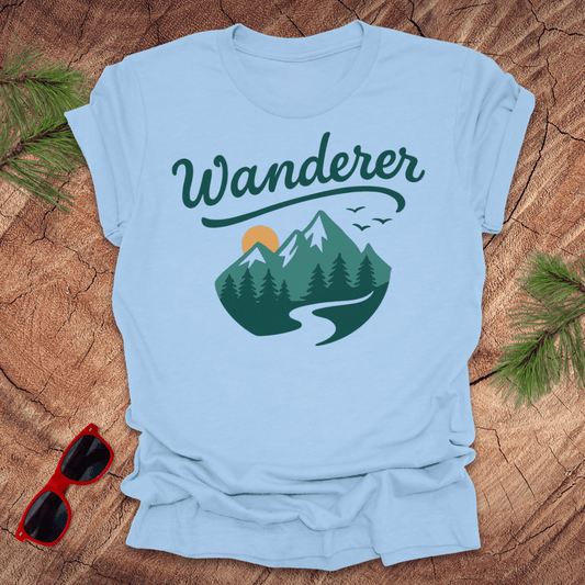 A blue crew neck t-shirt with a graphic print of mountains and the word 'Wanderer' on the front, displayed on a wooden surface with pine needles and a pair of sunglasses to the side.