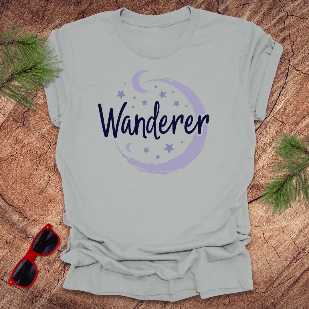 silver crew neck t-shirt with a moon and stars design, and the word 'Wanderer' written across the chest. The shirt is presented on a wooden background with pine needles and a pair of sunglasses beside it.