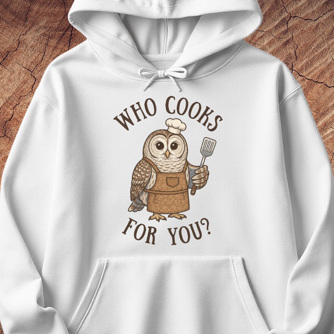 A white pullover hoodie featuring a cartoon owl wearing a brown apron and chef's hat, holding a spatula, with the text 'WHO COOKS FOR YOU?' in brown above and below. The hoodie is laid flat on a textured wood background.