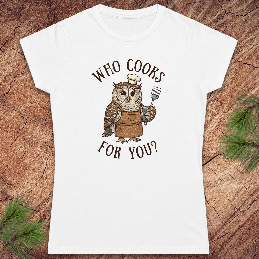 A white short-sleeve crewneck t-shirt featuring a cartoon owl dressed as a chef with the text 'WHO COOKS FOR YOU?' printed on the front, displayed flat on a rustic wood background with pine needles.