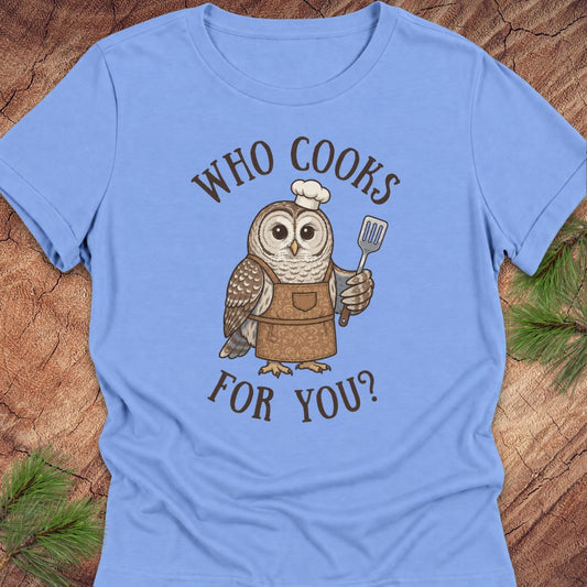 A light blue short-sleeve t-shirt laid flat on a rustic wood background with pine branches in the corners. The shirt features a cartoon owl wearing a brown apron and white chef's hat, holding a spatula, with the text 'WHO COOKS FOR YOU?' in brown above and below the graphic.