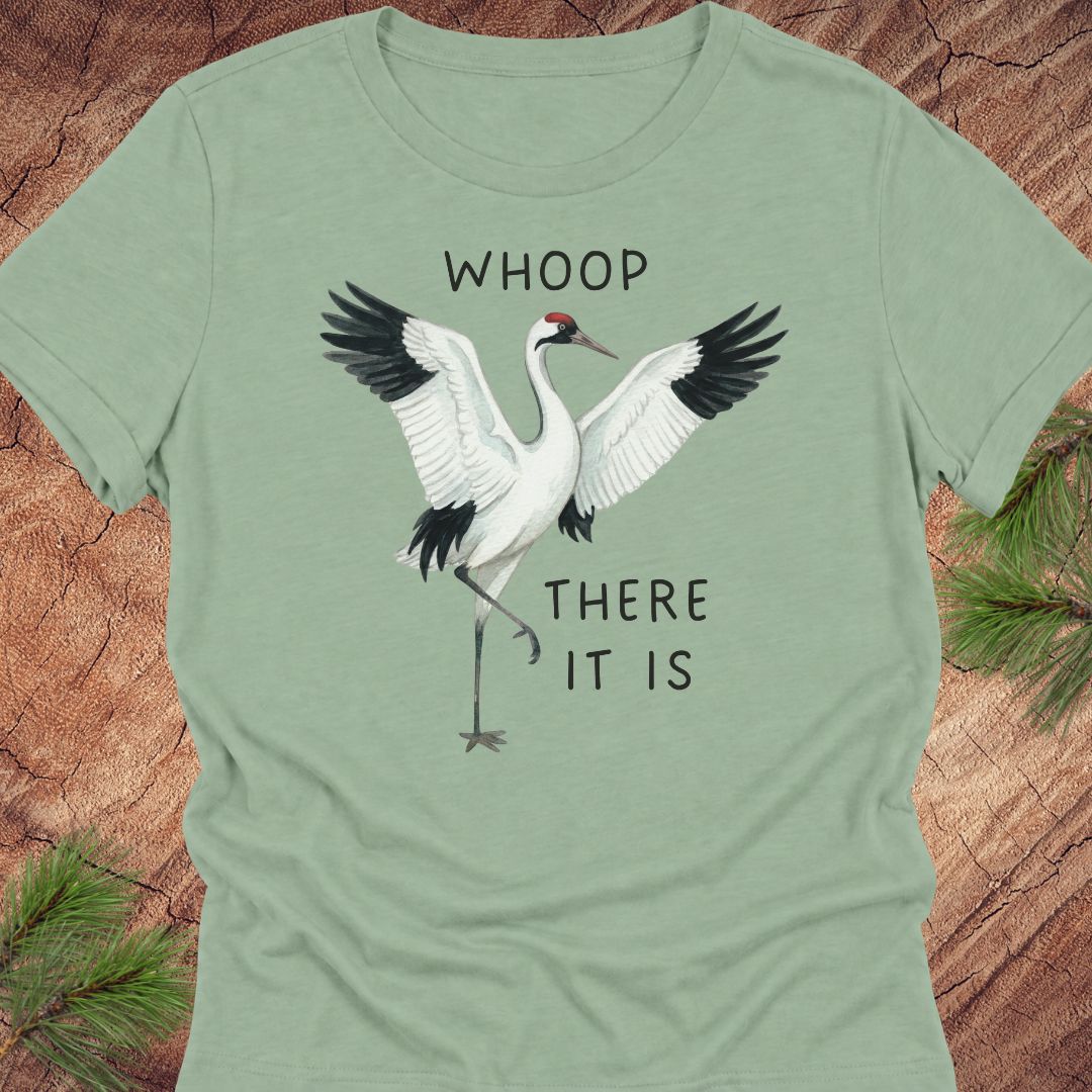 A sage green short-sleeve t-shirt featuring a graphic of a white crane with black wings and the text 'WHOOP THERE IT IS' printed on the front, laid flat on a rustic wood background with pine branches.