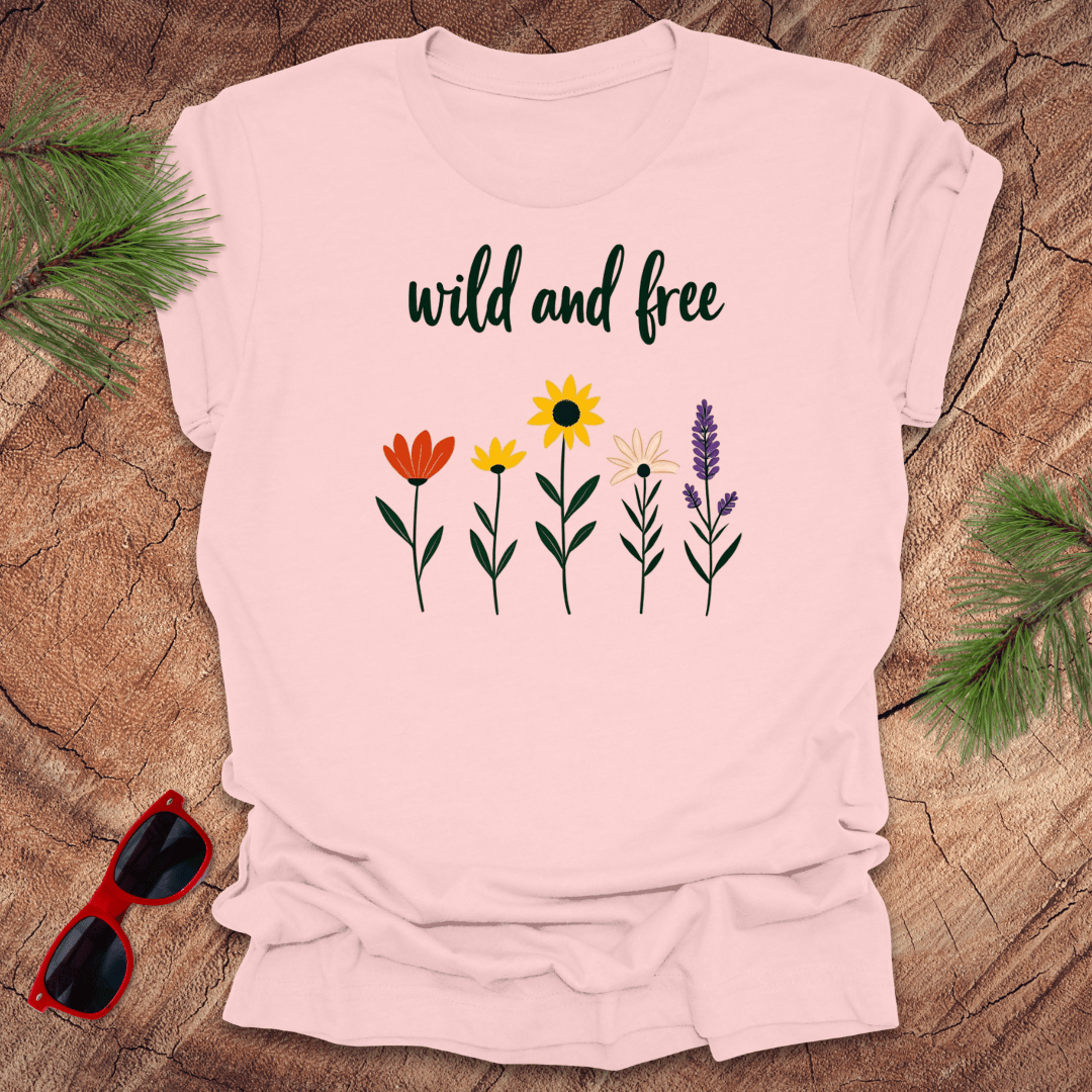 A light pink t-shirt with a graphic of various flowers and the text 'wild and free' displayed on a wooden surface with sunglasses to the side.