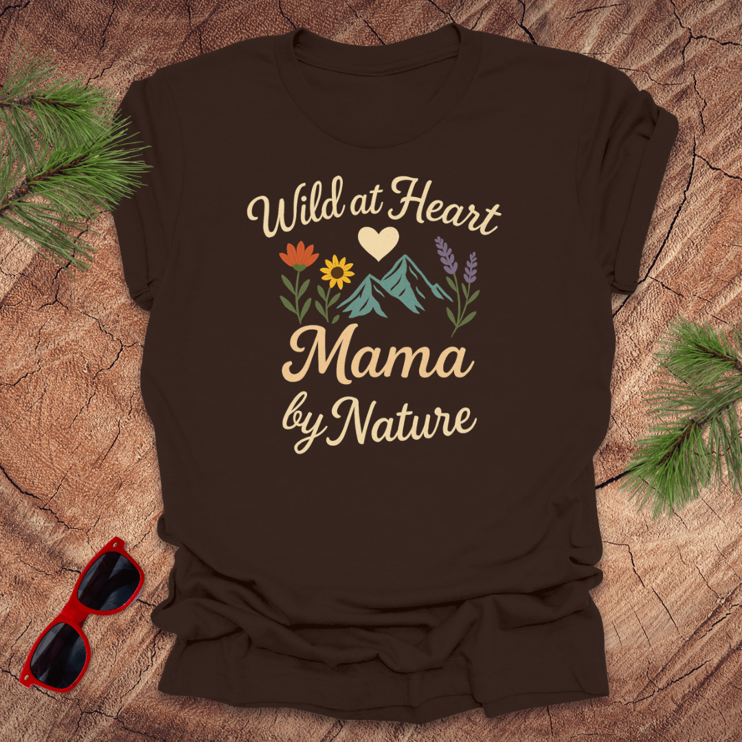 A brown t-shirt with a graphic that includes mountains, flowers, and the text 'Wild at Heart, Mama by Nature'. The shirt is on a wood background with pine needles and a pair of sunglasses.