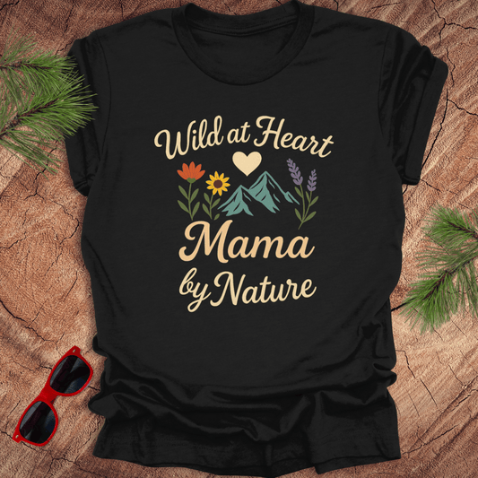 A black t-shirt with a graphic that includes mountains, flowers, and the text 'Wild at Heart, Mama by Nature'. The shirt is on a wood background with pine needles and a pair of sunglasses.