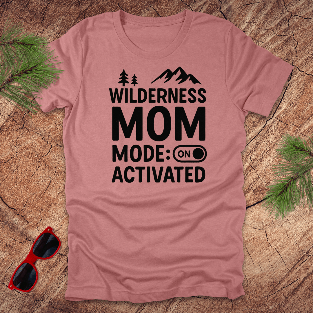 mauve t-shirt with the text 'Wilderness MOM Mode: Activated' printed in black, accompanied by silhouettes of trees and mountains. The shirt is presented on a wooden surface with a pair of red sunglasses.