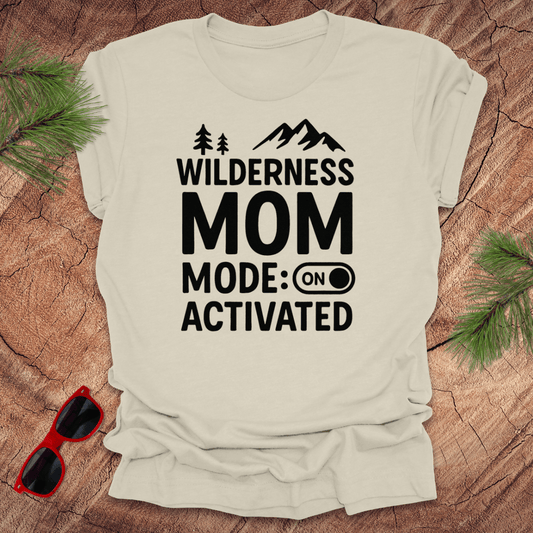 beige t-shirt with the text 'Wilderness MOM Mode: Activated' printed in black, accompanied by silhouettes of trees and mountains. The shirt is presented on a wooden surface with a pair of red sunglasses.