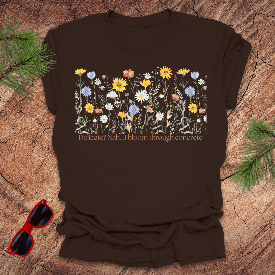 Brown t-shirt with floral design and text "delicate? nah. i bloom through concrete.' on a wooden surface.