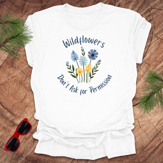 White t-shirt with floral design and text 'Wildflowers don't ask for permission' on a wooden surface with sunglasses and greenery.