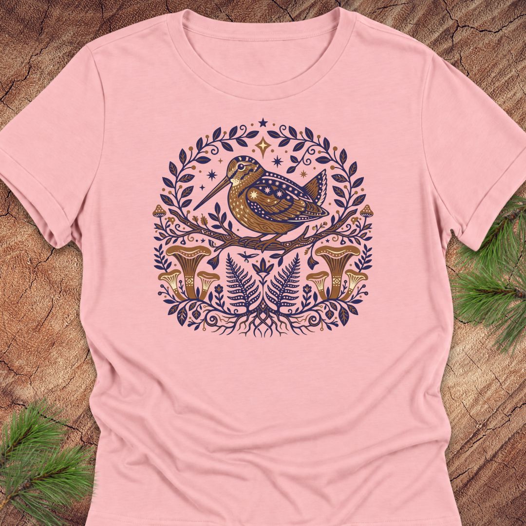 A light pink short-sleeve crewneck t-shirt featuring a detailed graphic of a brown bird with blue spots, surrounded by a circular wreath of dark blue leaves and ferns, with brown mushrooms at the bottom. The t-shirt is laid flat on a rustic wood background with pine needles in the corners.