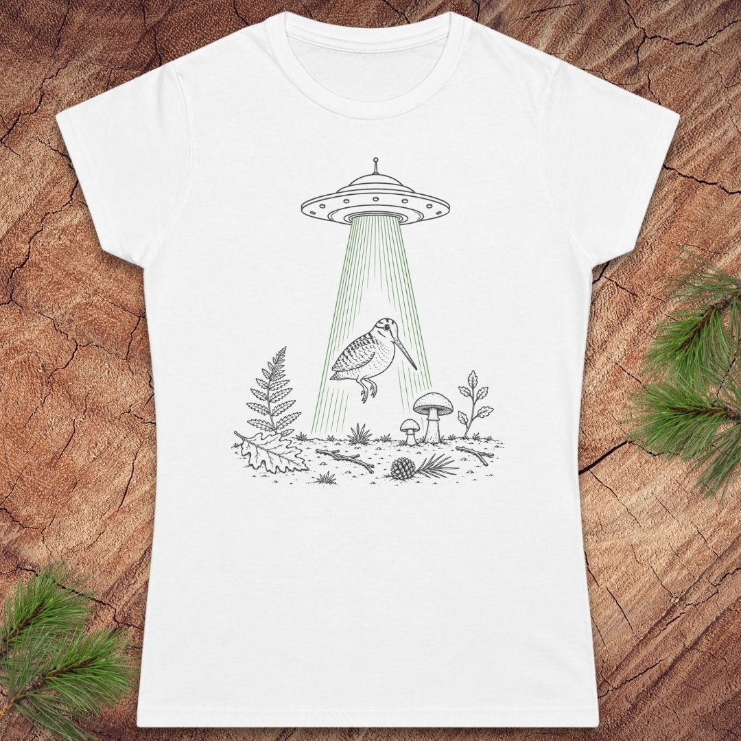 A white short-sleeve crewneck t-shirt featuring a black line art graphic of a UFO with a green beam of light shining down onto a bird, ferns, and mushrooms, laid flat on a rustic wood background with pine branches in the corners.
