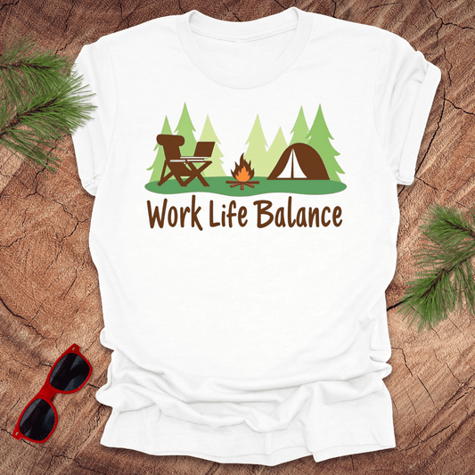 A white t-shirt with a graphic print of a camping scene including a chair, tent, fire, and trees, with the text 'Work Life Balance'. There are sunglasses and pine branches on the wooden surface around the shirt.