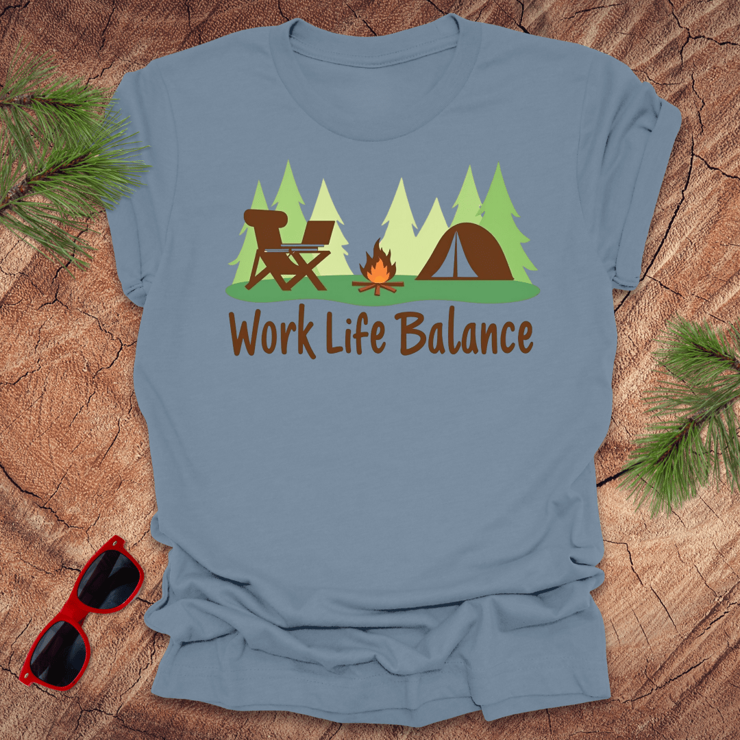 A blue t-shirt with a graphic print of a camping scene including a chair, tent, fire, and trees, with the text 'Work Life Balance'. There are sunglasses and pine branches on the wooden surface around the shirt.
