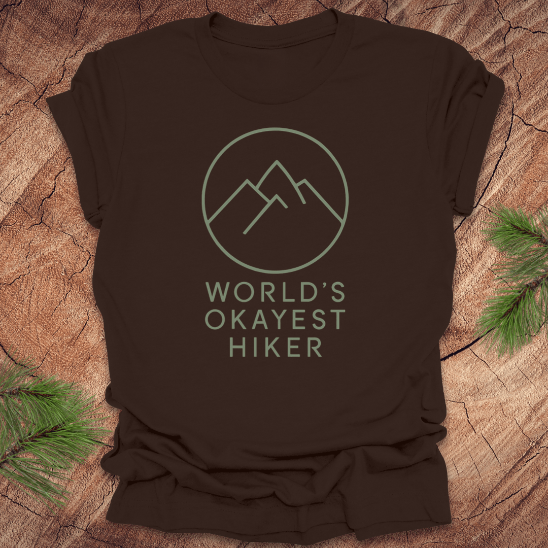 Brown t-shirt with 'World's Okayest Hiker' graphic on a wooden surface with pine branches.