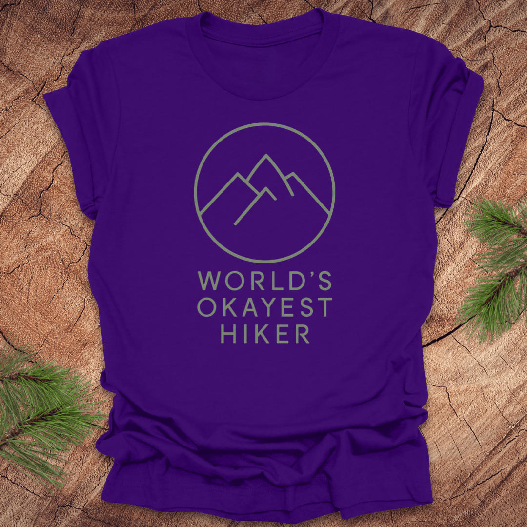 Purple t-shirt with mountain graphic and 'World's Okayest Hiker' text on a wooden surface.
