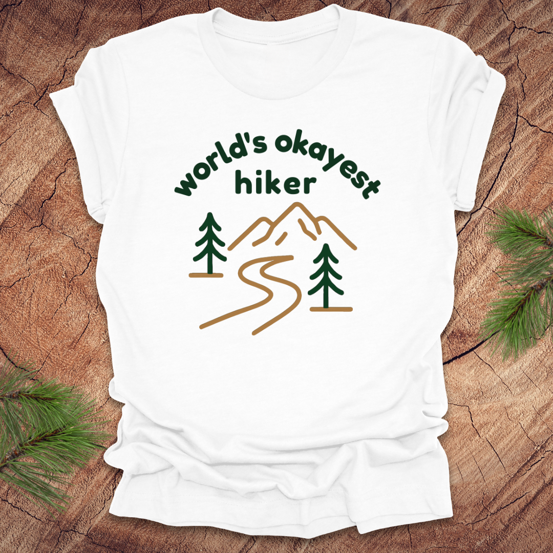 White t-shirt with 'world's okayest hiker' graphic on a wooden surface with pine branches.