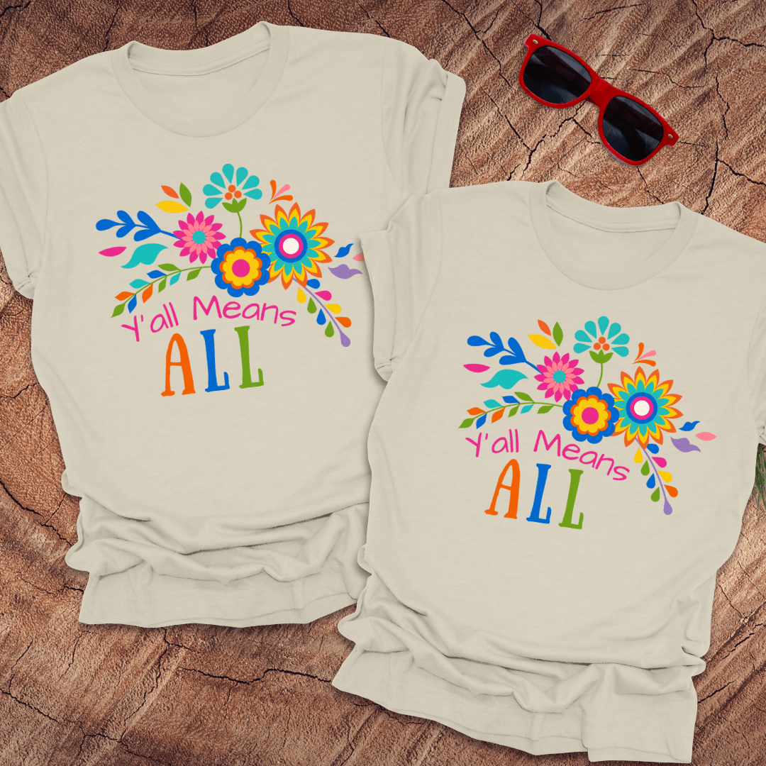 Two light-colored t-shirts with colorful floral designs and text "Y'all Means All' on a wooden surface.
