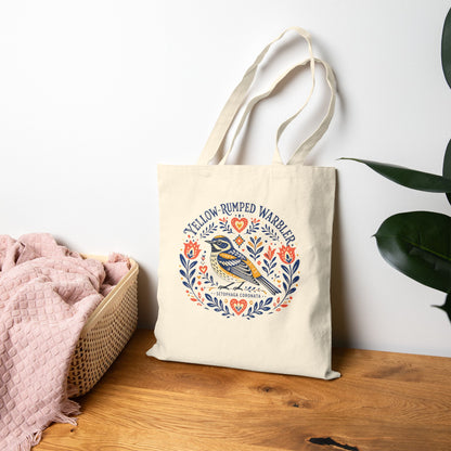 Canvas tote bag with a folk art yellow-rumped warbler design on a wood table.