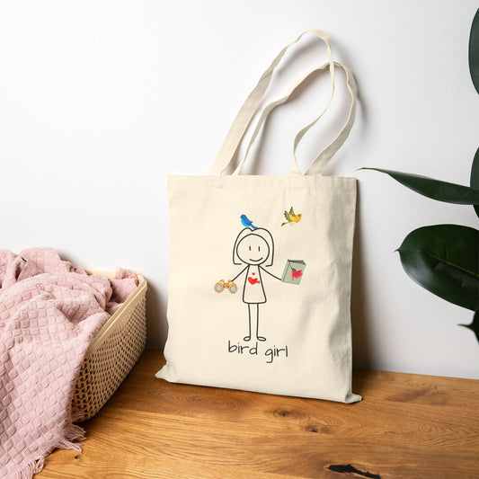 Tote bag with 'bird girl' design on a wooden surface next to a basket of pink items.