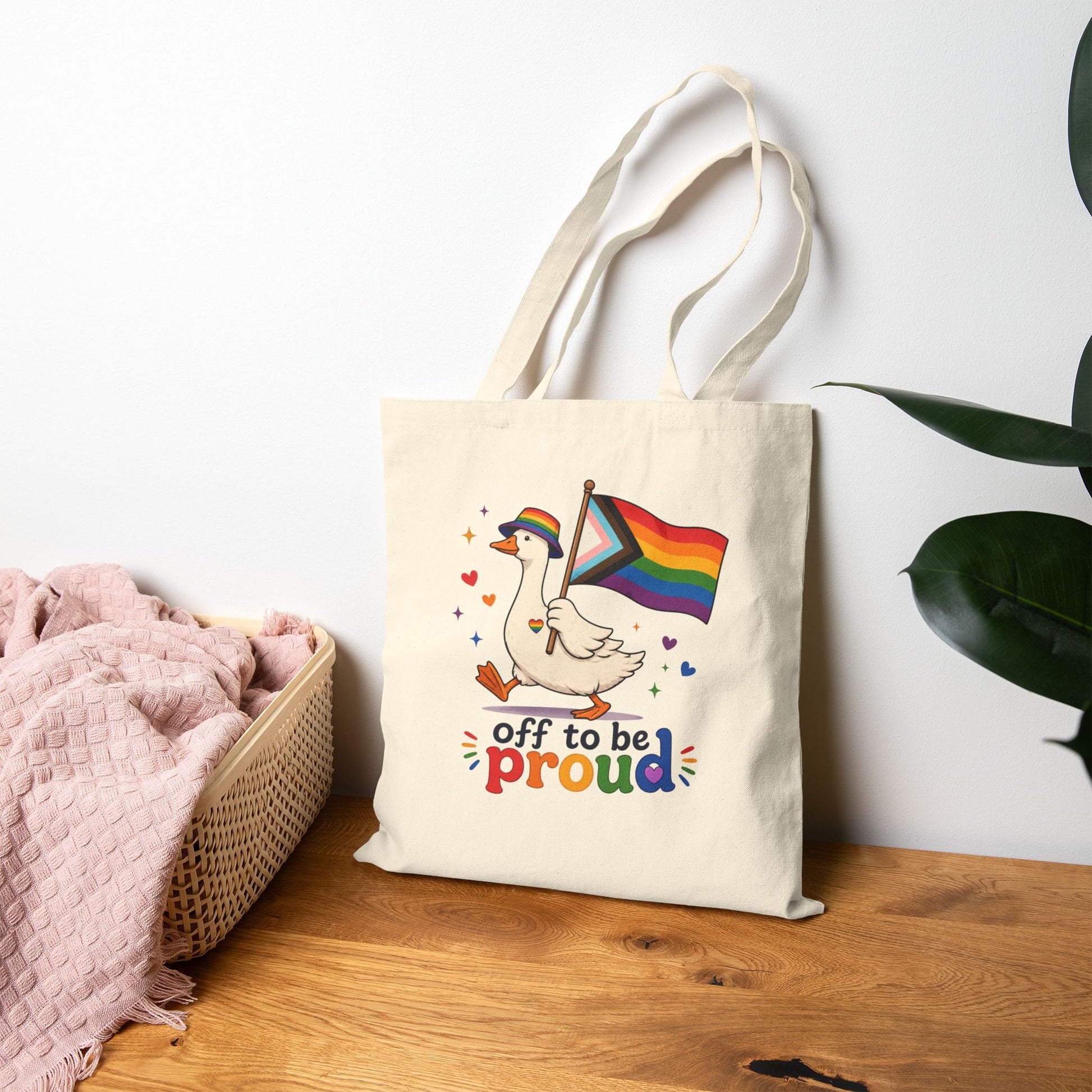 Tote bag with a colorful design featuring a goose holding a rainbow flag and text 'off to be proud' on a wooden surface.