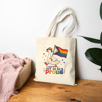 Tote bag with a colorful design featuring a goose holding a rainbow flag and text 'off to be proud' on a wooden surface.