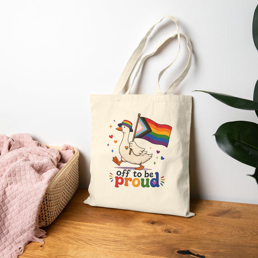 Tote bag with a colorful design featuring a goose holding a rainbow flag and text 'off to be proud' on a wooden surface.