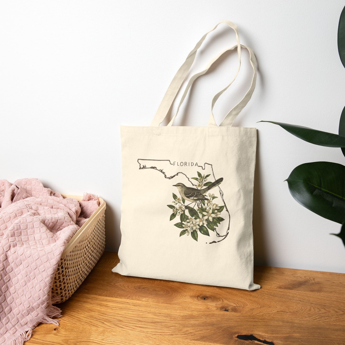 Tote bag with Florida bird design on a wooden surface next to a basket and plant