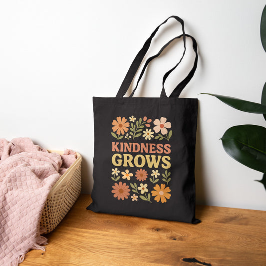 Black tote bag with 'Kindness grows' text on a wooden surface with a plant and pink blanket.