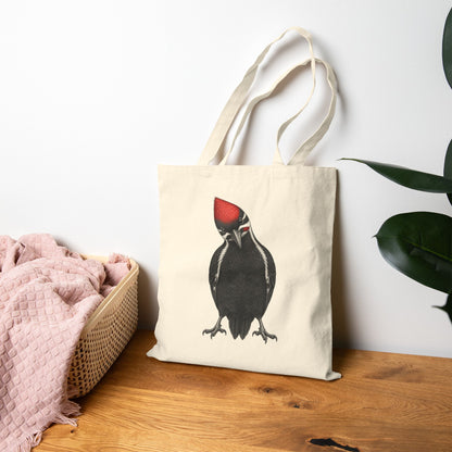 Tote bag with a woodpecker design on a wooden surface next to a pink blanket and plant.