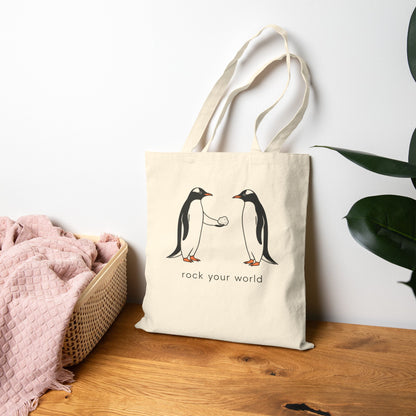 Beige canvas tote bag with a penguin design and text 'rock your world' on a table.