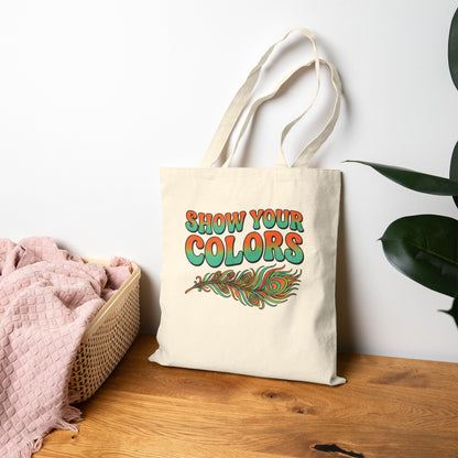Tote bag with 'Show Your Colors' text and  peacock feather design on a wooden surface.