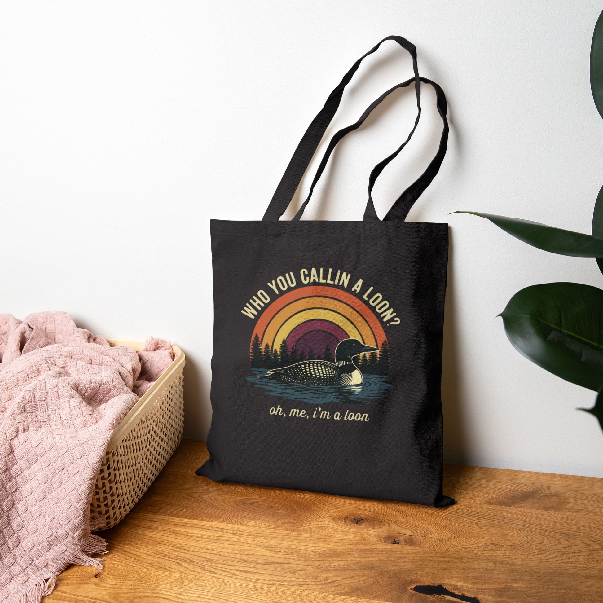 Black tote bag with loon design and text on a wooden surface with a plant in the background