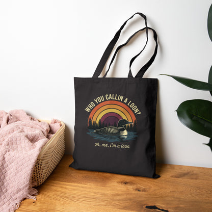 Black tote bag with loon design and text on a wooden surface with a plant in the background
