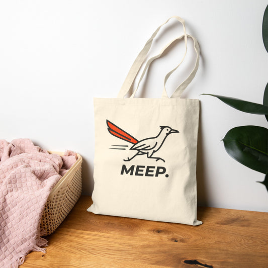 Beige tote bag with meep roadrunner design on a wooden surface with a white wall and plant in the background