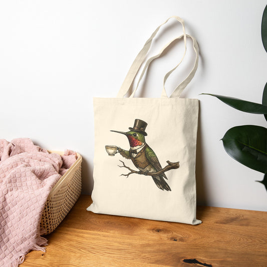 A canvas tote bag with a hummingbird in a suit and top hate, with tea design, on a table.