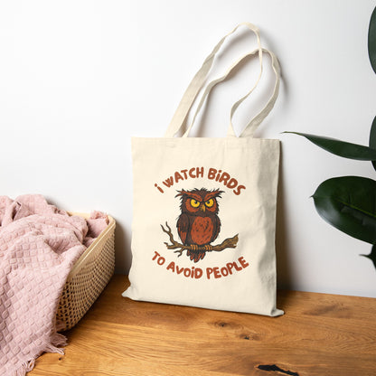 A canvas tote bag with owl design and text, 'i watch birds to avoid people' on a table. 