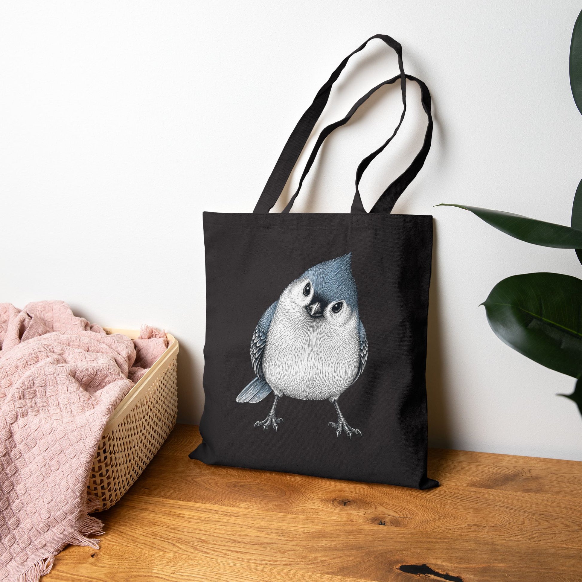 Black tote bag with a titmouse design on a wooden surface with a white wall and plant in the background