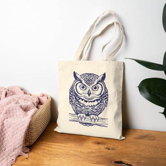 Tote bag with a linocut owl design on a wooden surface next to a pink blanket and plant.