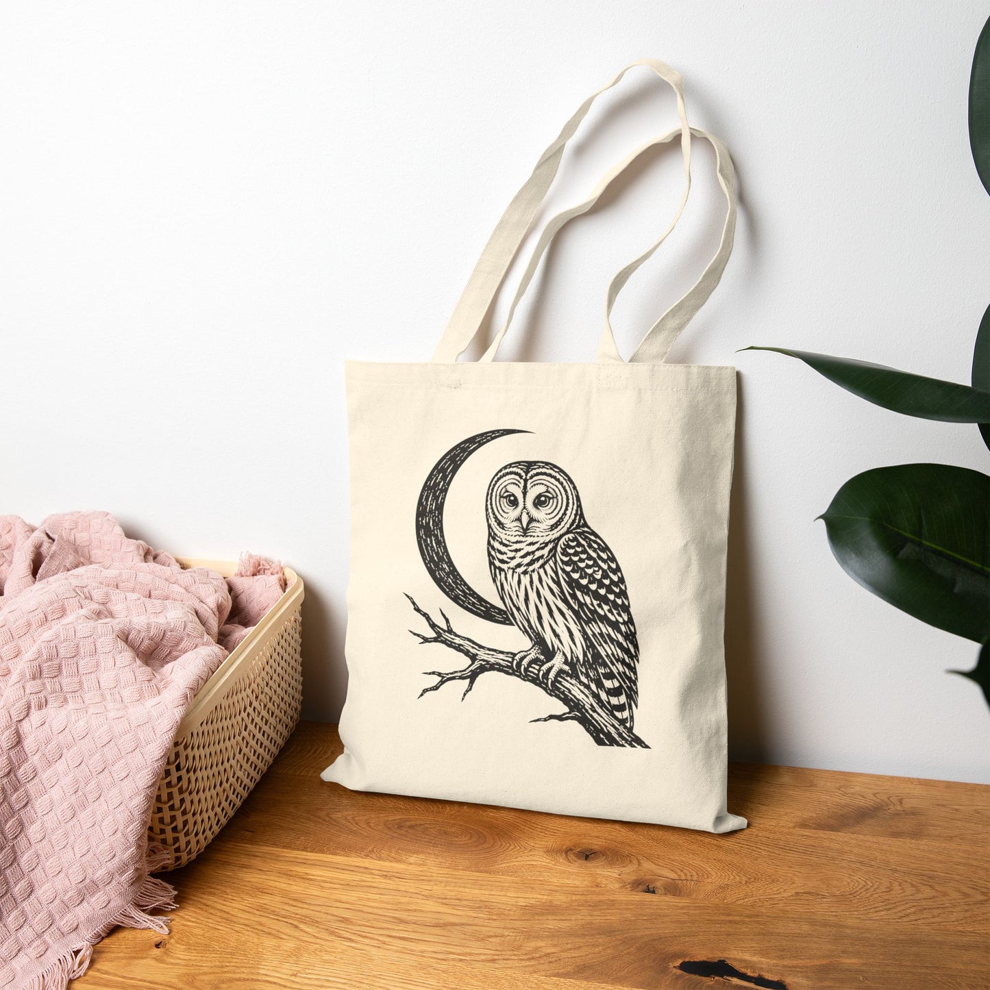 An off-white canvas tote bag with a black line art illustration of an owl perched on a crescent moon branch, displayed on a wooden table next to a pink blanket and a green plant against a white wall.