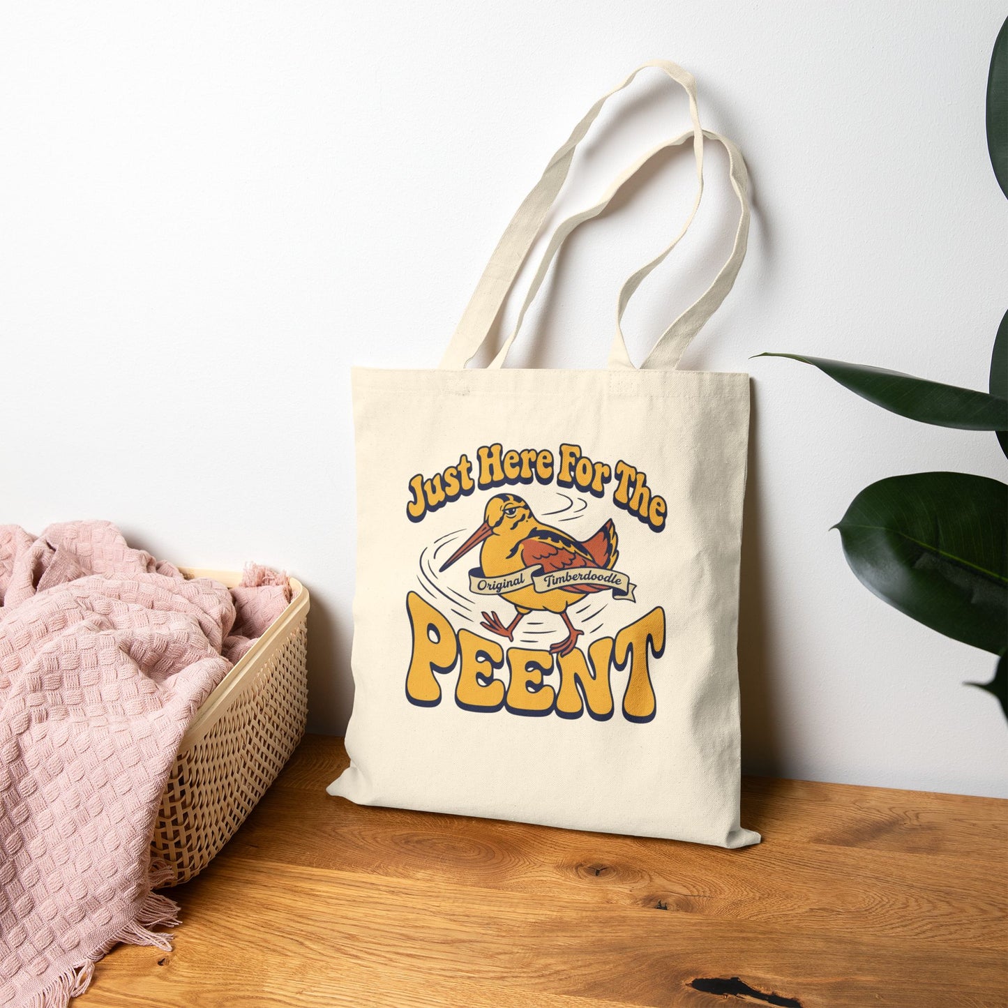 Tote bag with 'Just Here For The Peent' design on a wooden surface with a white wall and plant in the background