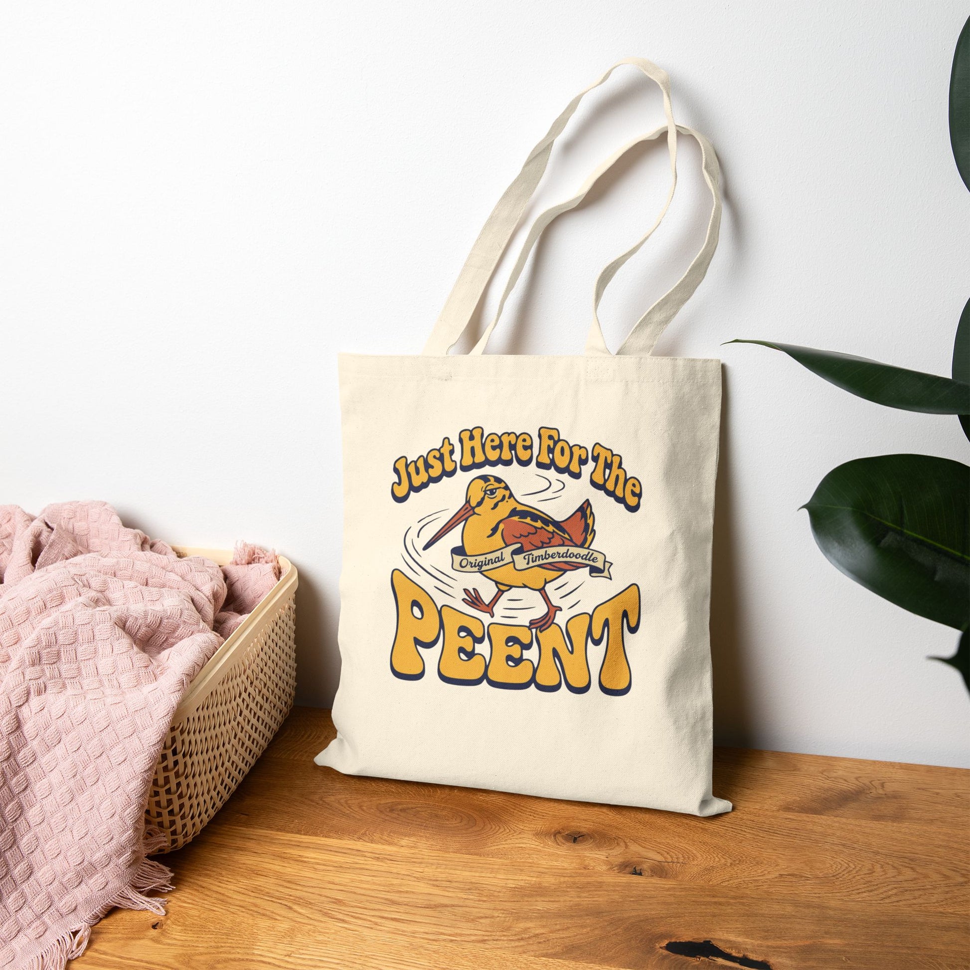 Tote bag with 'Just Here For The Peent' design on a wooden surface with a white wall and plant in the background