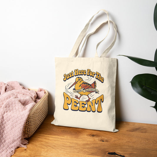 Tote bag with 'Just Here For The Peent' design on a wooden surface with a white wall and plant in the background