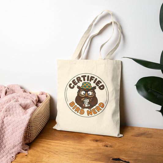 Tote bag with 'Certified Bird Nerd' design on a wooden surface with a white wall and plant in the background.