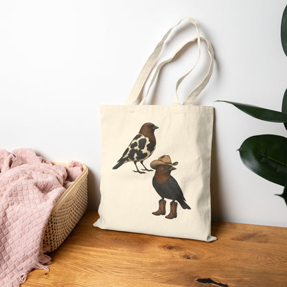 An off-white canvas tote bag with two illustrated birds printed on the front, standing on a wooden surface against a white wall. A pink textured blanket in a woven basket is on the left, and a green plant leaf is on the right.