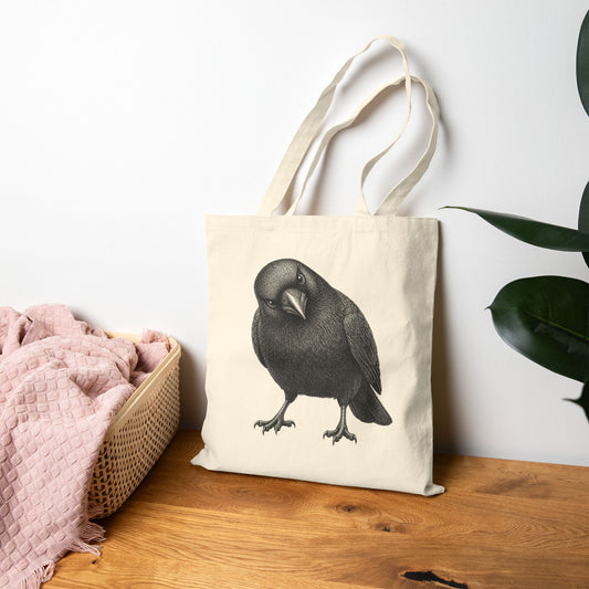Tote bag with a crow design on a wooden surface next to a pink blanket and plant.