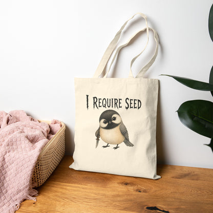 A cream canvas tote bag with long handles, featuring a black text graphic that reads 'I REQUIRE SEED' above a small chickadee illustration. The bag is styled on a wooden table next to a pink blanket in a basket and a green plant, against a white wall.